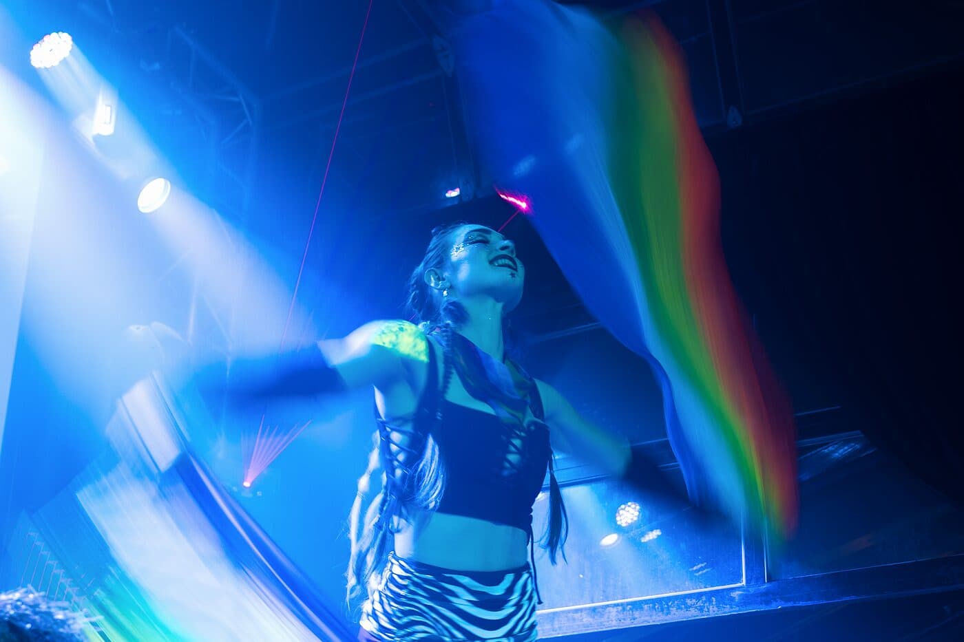 A performer under blue stage light, a rainbow flag sweeping through the frame