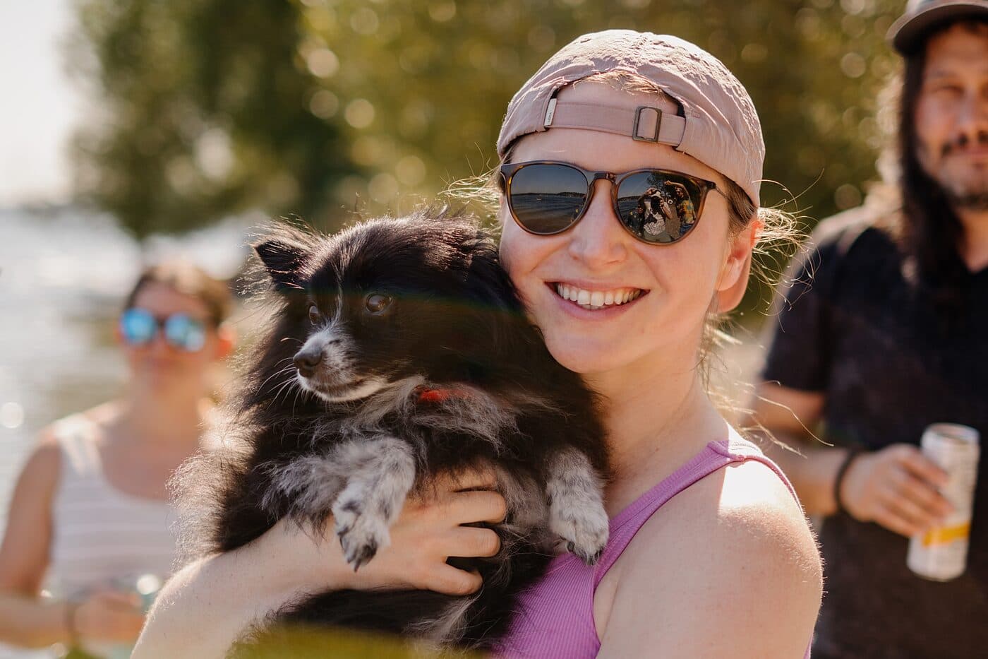 A woman holding a small dog at a sunlit lakeside gathering
